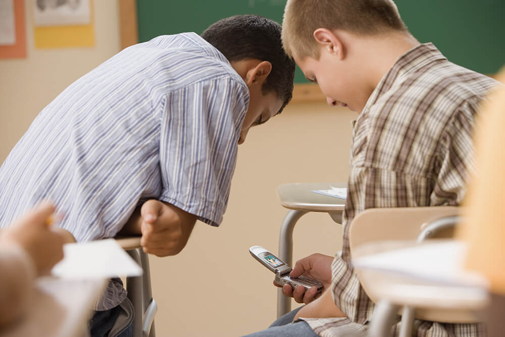 two young male students laugh in class on a flip phone exhibiting teen media use