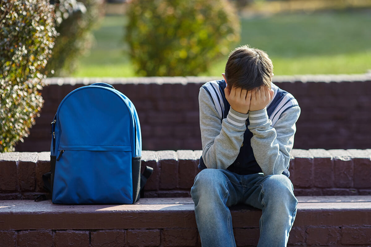 teen boy with hands over face sitting next to backpack showing signs of drug use