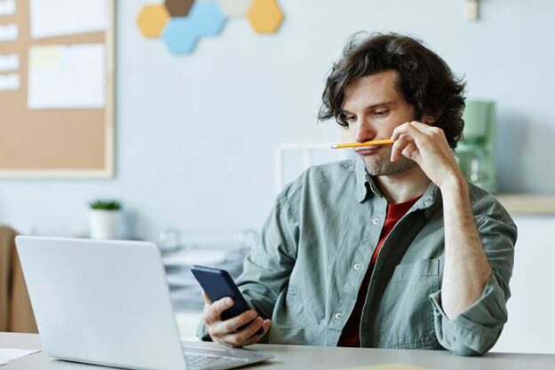 a teen balances a pencil on his upper lip while playing on his phone instead of doing schoolwork