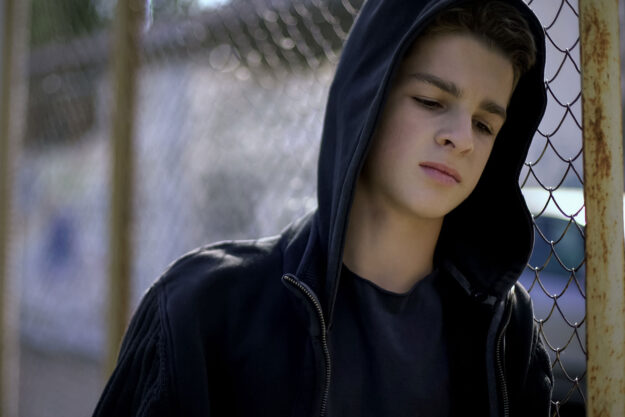 a delinquent teen with his hood up stands against a fence